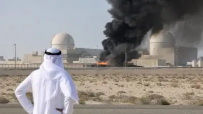 Ein Mann in traditioneller arabischer Kleidung steht vor einem Kernkraftwerk mit schwarzem Rauch und Feuer.