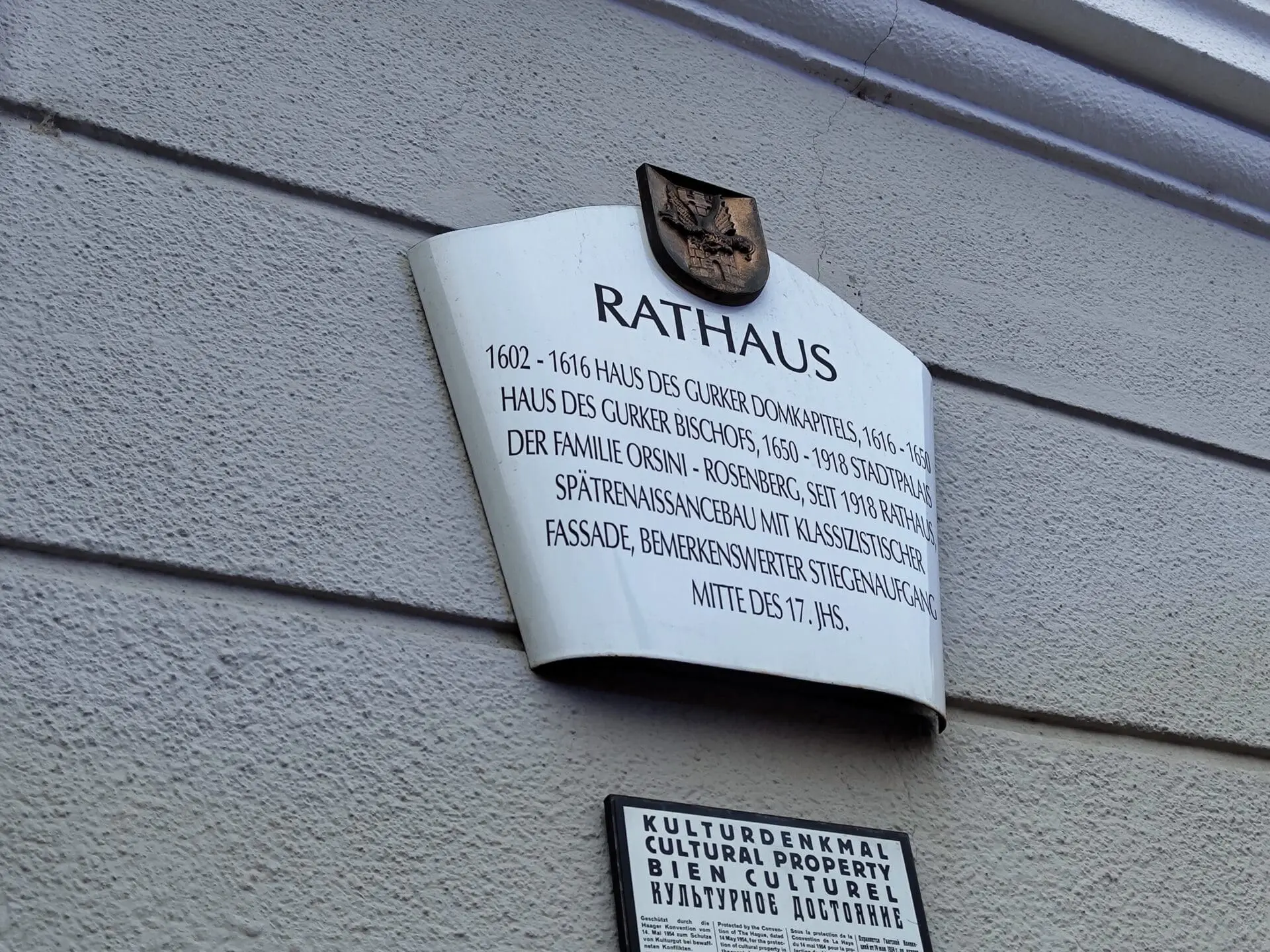 Weißes Schild mit Aufschrift Rathaus und historischen Daten an grauer Hauswand
