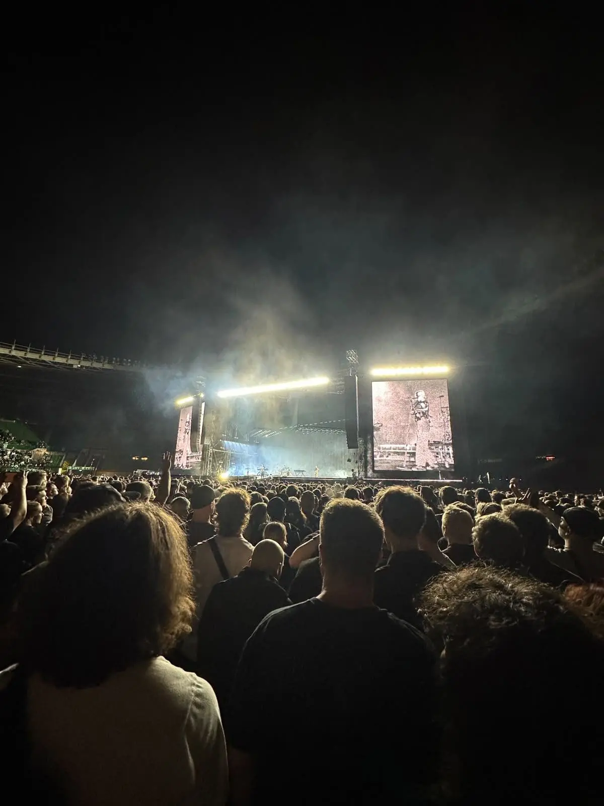 Blick von hinten auf eine dichte Menschenmenge bei einem nächtlichen Konzert in einem Stadion mit beleuchteter Bühne.