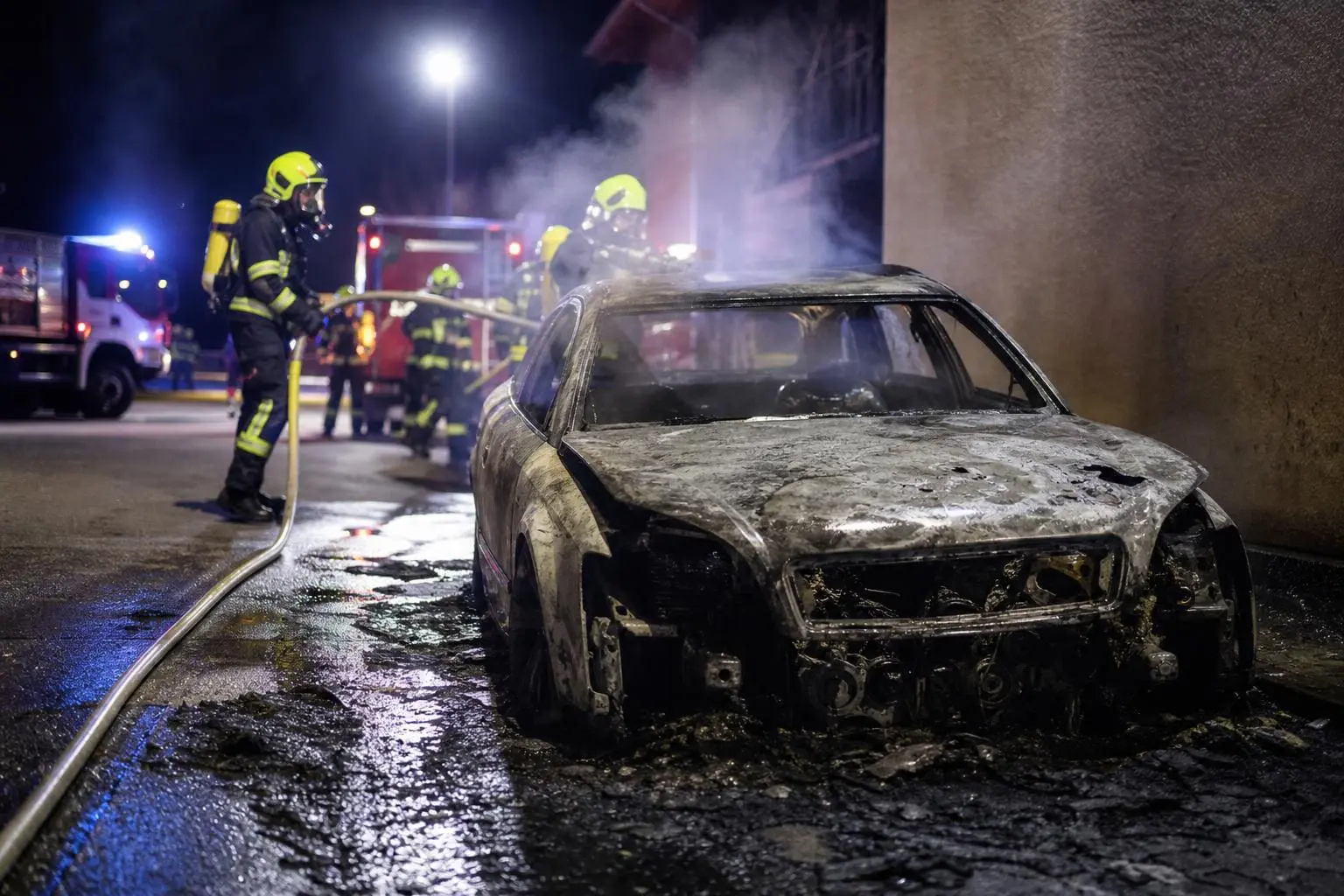 Völlig ausgebrannter silberner Pkw mit Feuerwehrleuten und Löschschläuchen bei Nacht