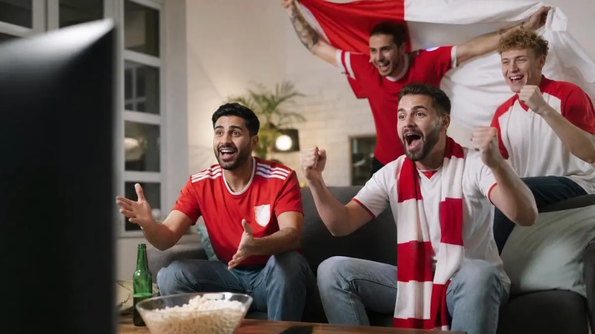 Drei Männer in roten Fußballtrikots jubeln vor einem Fernseher im Wohnzimmer