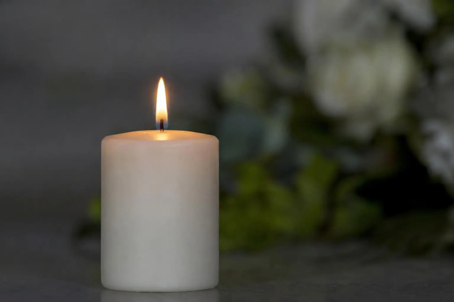 Brennende weiße Kerze im Vordergrund, unscharfe Blumen im Hintergrund