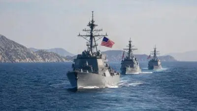 Drei graue US-Zerstörer mit wehenden amerikanischen Flaggen fahren in Formation auf dem Meer.