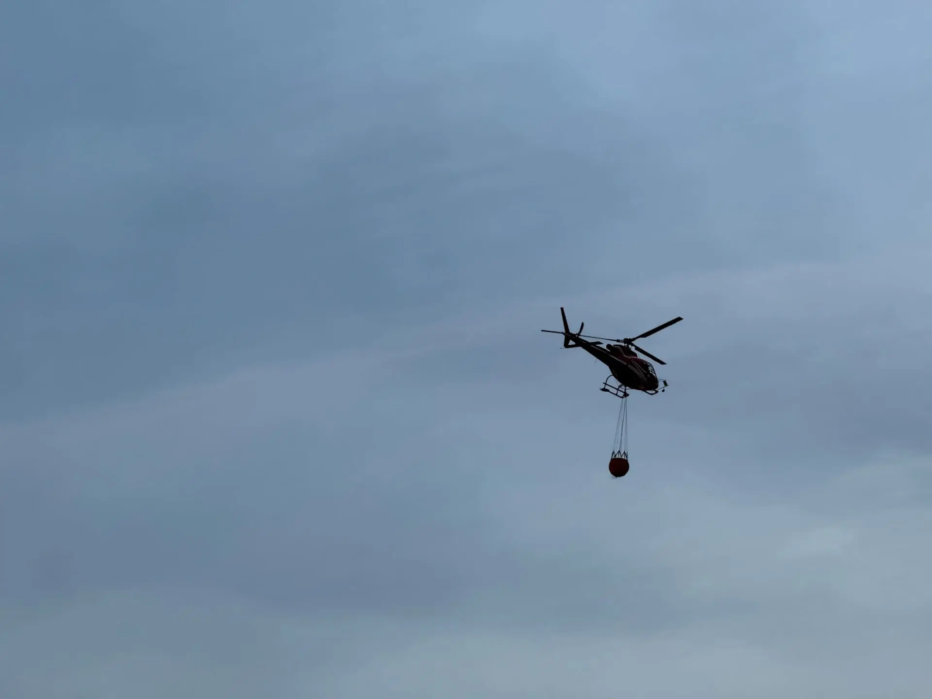 Ein Hubschrauber mit einem roten Behälter fliegt vor einem bewölkten Himmel.