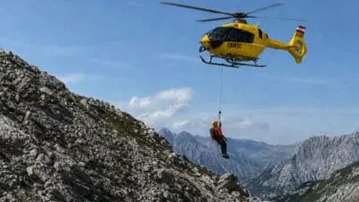Ein gelber Rettungshubschrauber mit einem am Seil hängenden Retter über einem steinigen Berg in den Alpen.