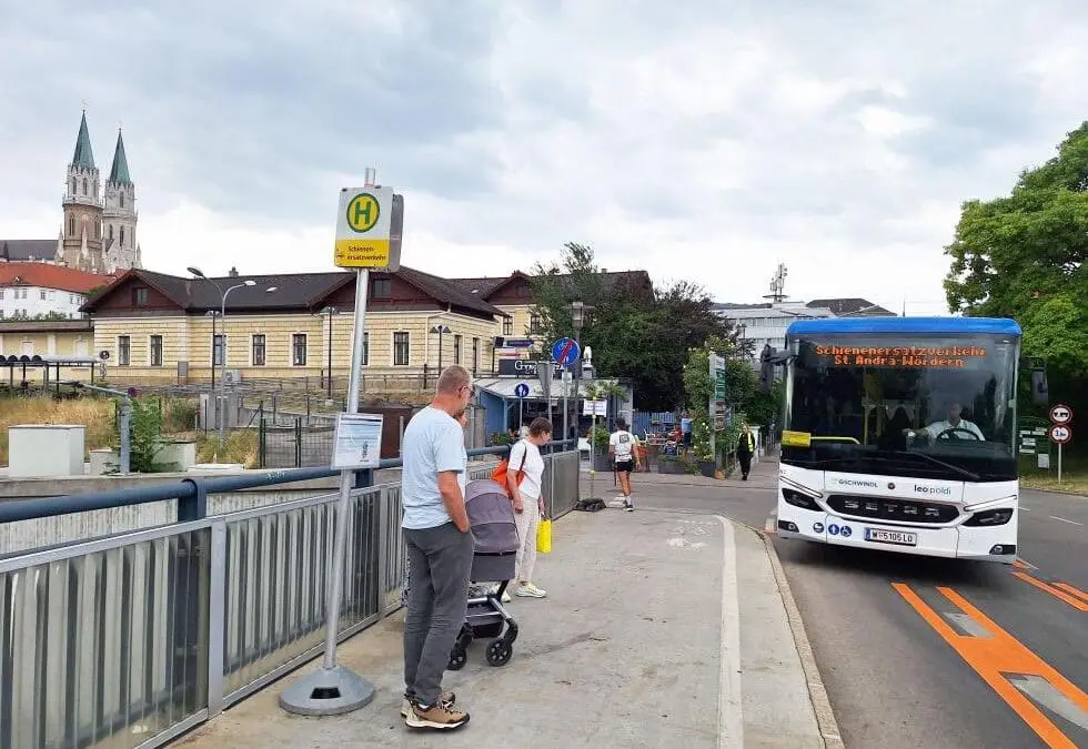 Weiß-blauer Bus mit Aufschrift Schienenersatzverkehr St. Andrä-Wördern fährt an Bushaltestelle mit wartenden Personen und Kinderwagen vorbei