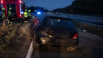 Feuerwehrleute in Warnwesten neben rotem Einsatzfahrzeug und beschädigtem schwarzen Pkw auf nächtlicher Autobahn