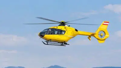 Gelber Hubschrauber fliegt vor blauem Himmel über einer Hügellandschaft.