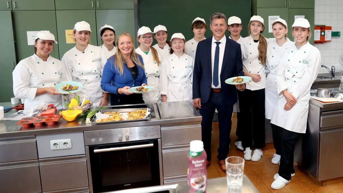 Landeshauptmann Daniel Fellner und Schülerinnen und Schüler präsentieren Speisen in einer Lehrküche.