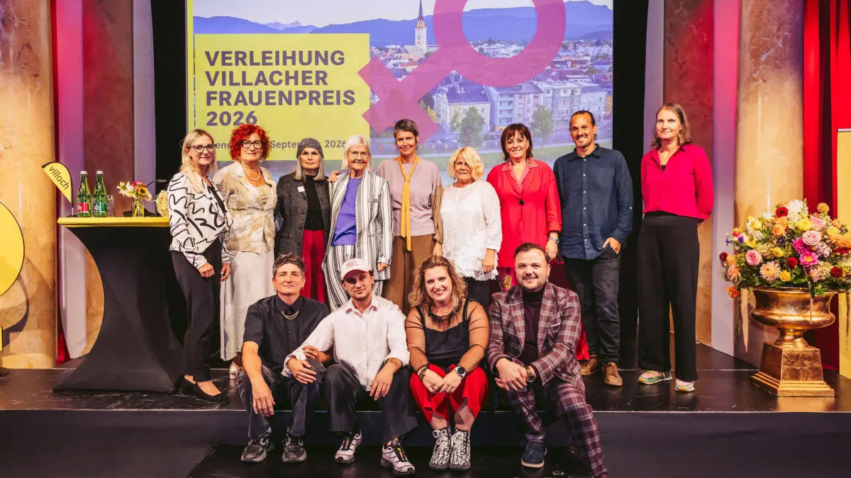 Eine Gruppe steht und sitzt auf einer Bühne vor der Tafel zum Villacher Frauenpreis 2026.