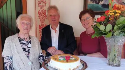 Ältere Frau, Mann im Anzug und Frau mit Brille sitzen an einem Tisch mit Torte.