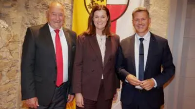 Drei Personen in formeller Kleidung stehen vor einer Fahne mit dem Wappen Kärntens.