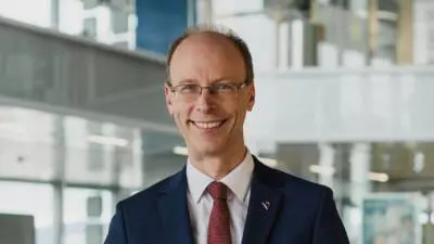 Lächelnder Mann in einem blauen Anzug mit roter Krawatte vor großen Fenstern.