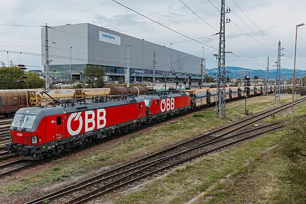 Rote ÖBB-Elektrolokomotive zieht Güterzug vor voestalpine-Gebäude auf Bahngleisen.