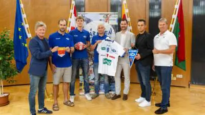 Männer stehen nebeneinander in einer Veranstaltungshalle und präsentieren ein weißes Handballtrikot sowie einen blauen Wimpel.