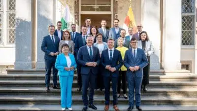 Gruppenporträt von zwanzig Frauen und Männern in Anzügen auf Steinstufen vor einem Gebäude mit Flaggen.