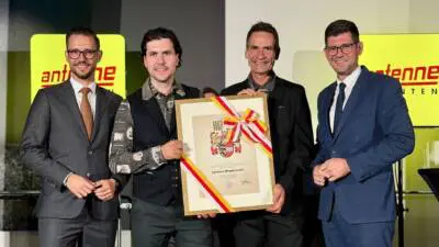Vier Männer in Anzügen präsentieren eine gerahmte Urkunde mit Wappen und rot-weiß-rotem Band.