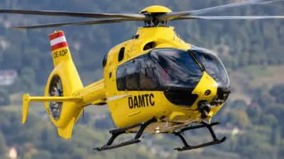 Ein gelber Rettungshubschrauber fliegt vor bewaldeten Bergen.