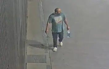 Ein Mann geht neben einer Wand über den Gehweg und trägt eine Flasche.