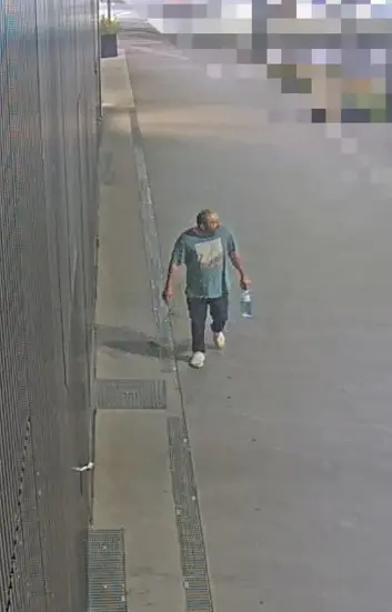 Ein Mann geht neben einer Wand über den Gehweg und trägt eine Flasche.