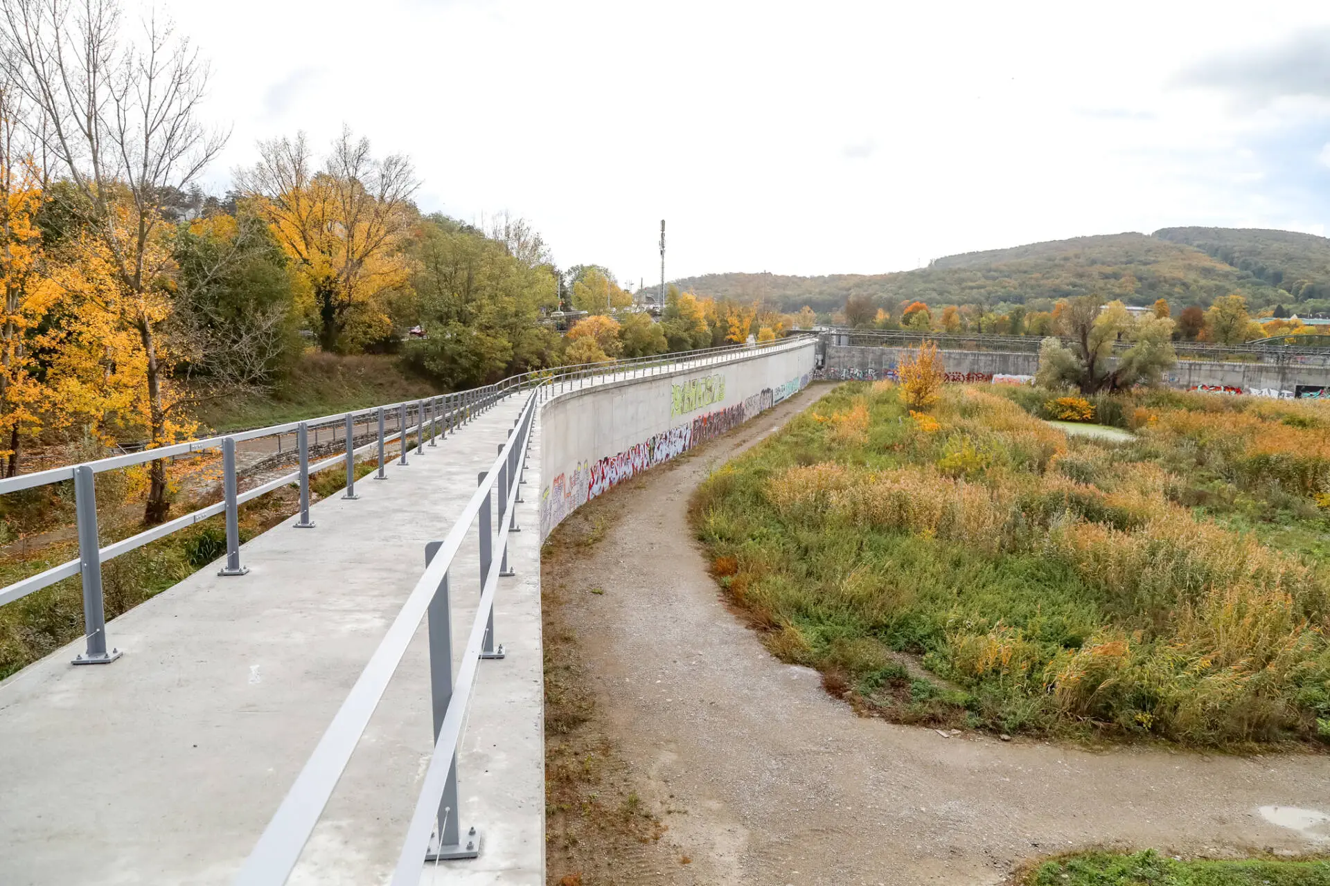 Betonmauer mit Metallgeländer neben einem begrünten, trockenen Becken im Herbst.