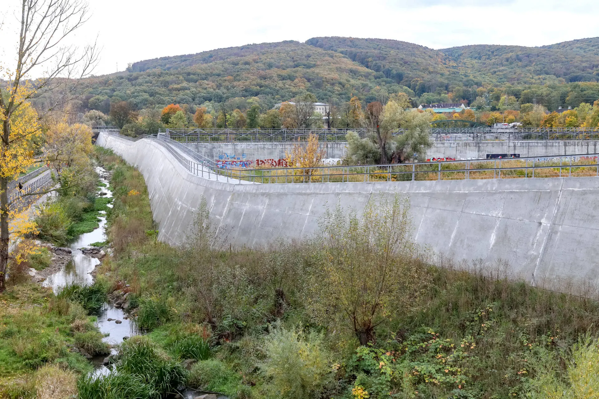 Hohe Betonmauern säumen einen begrünten Wasserlauf vor bewaldeten Hügeln.