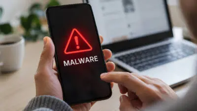 KI-generiertes Bild: Eine Hand hält ein Smartphone mit rotem Warnsymbol und dem Schriftzug „MALWARE“ vor einem Laptop.