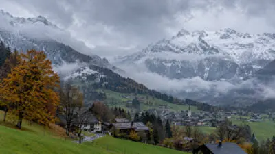 KI-generiertes Bild: Verschneite Berggipfel liegen über einem nebligen Tal mit grünen Wiesen, Häusern und herbstlich gefärbten Bäumen.