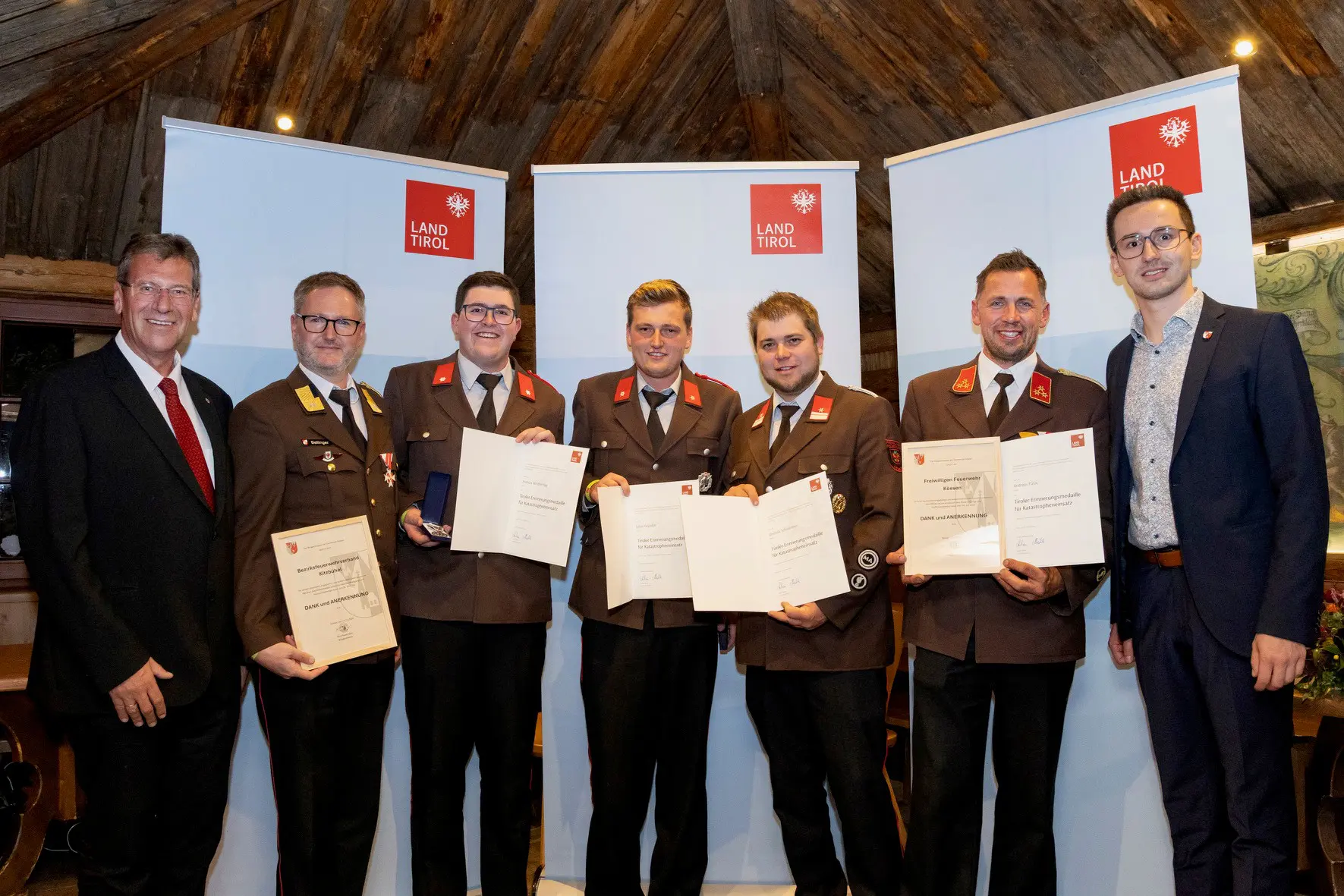 Personen in Feuerwehruniformen und Anzügen halten Urkunden und Medaillen.