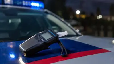 KI-Symbolbild: Schwarzes Atemalkoholmessgerät auf einer blau-weiß-roten Motorhaube vor blauem Blaulicht