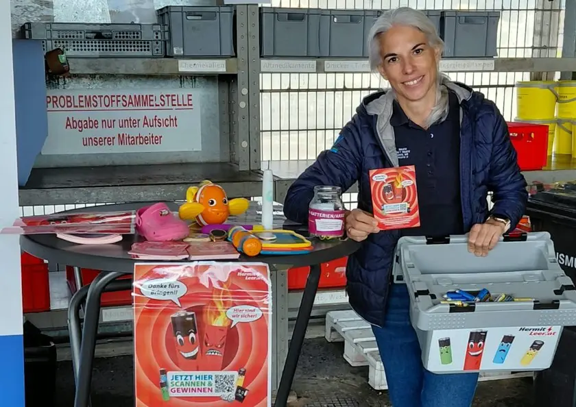 Eine Frau zeigt eine Akku-Infokarte neben Spielzeug und einer Sammelbox mit Batterien.
