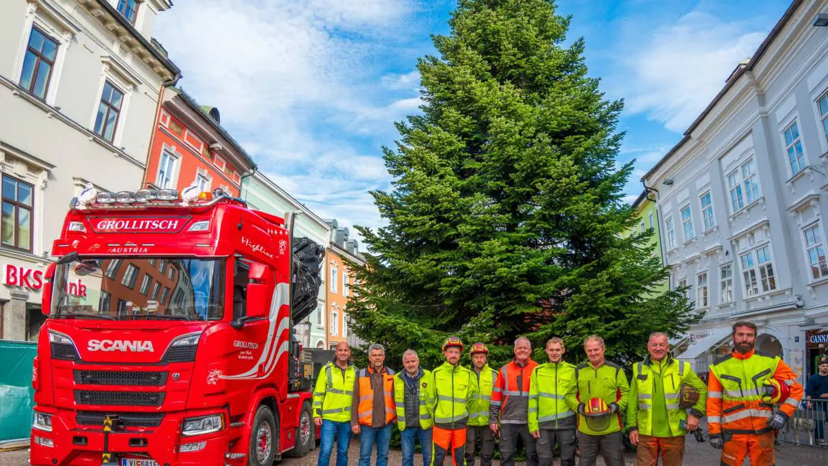Der Christbaum steht neben einem roten Lkw und Mitarbeitern auf dem Hauptplatz in Villach.