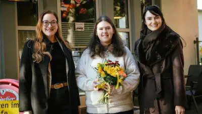 Drei Frauen stehen vor einem Café; die Frau in der Mitte hält einen bunten Blumenstrauß.