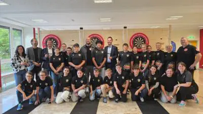 Schüler:innen und Erwachsene posieren vor drei Dartscheiben in einem Innenraum.