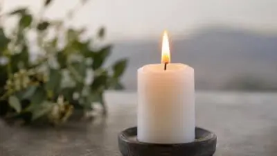 Eine brennende weiße Stumpenkerze steht in einem dunklen Kerzenhalter neben unscharfen Blumen.