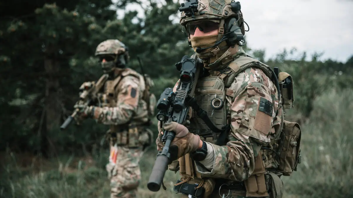 Ein Soldat in Tarnuniform hält ein Gewehr im Wald, ein weiterer steht dahinter.