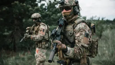Ein Soldat in Tarnuniform hält ein Gewehr im Wald, ein weiterer steht dahinter.