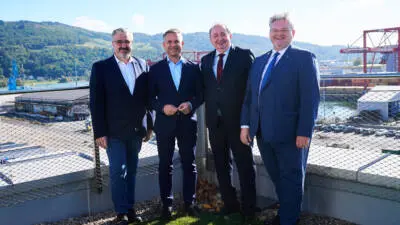 Männer in Anzügen stehen vor einer Hafenanlage mit Kränen und bewaldeten Hügeln.
