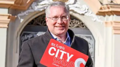 Ein Mann hält eine rote St.-Veiter City Card vor einem verzierten Hauseingang.