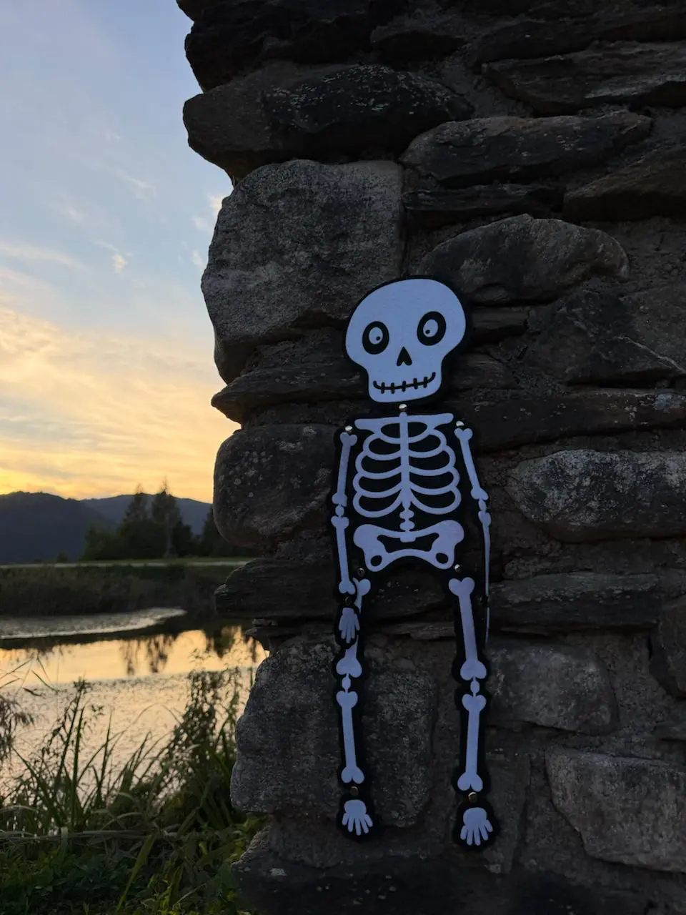 Ein Skelettmotiv hängt an einer Steinmauer vor einem See im Abendlicht.