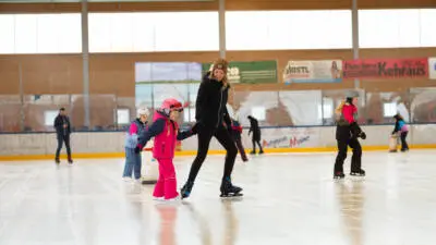 Eine Frau hilft einem Kind beim Eislaufen in einer überdachten Eishalle.