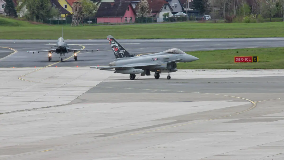 Eurofighter rollen über ein Flughafenvorfeld vor einer grünen Wiese.