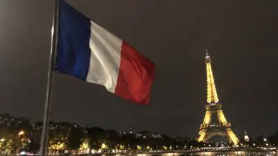KI-generiertes Bild: Eine französische Flagge weht neben einem beleuchteten Turm am Flussufer.