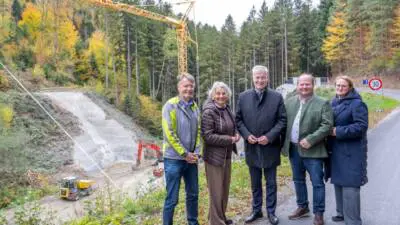 Personen stehen an einer Baustelle mit Bagger und Baukran am bewaldeten Hang.