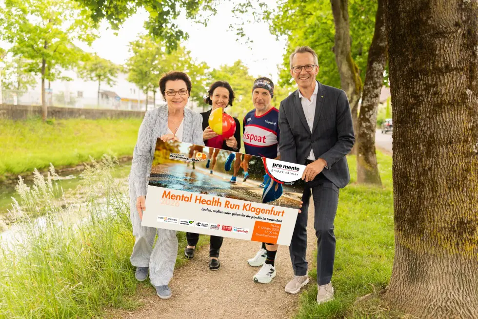 Erwachsene präsentieren ein Schild zum Mental Health Run Klagenfurt auf einem Weg im Grünen.