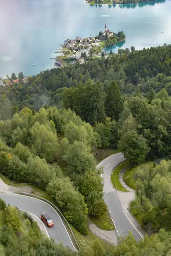 Luftaufnahme einer kurvigen Straße durch Wald mit See und Ortschaft im Hintergrund