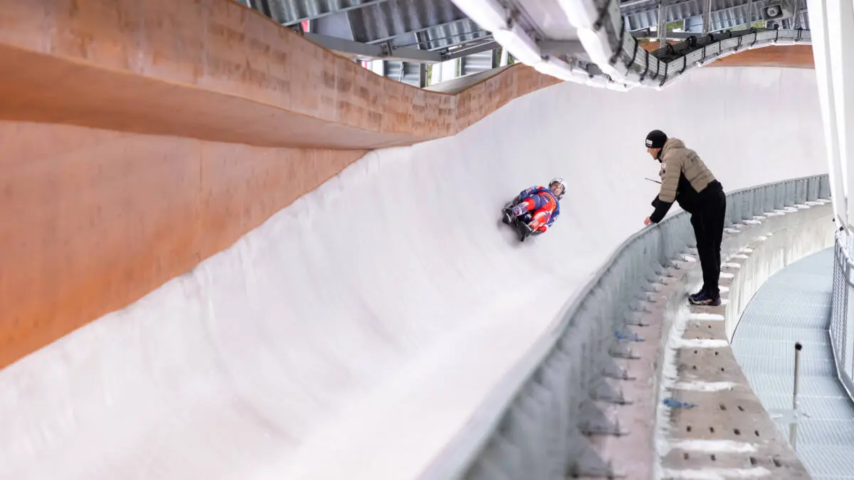 Ein Athlet fährt auf einem Eiskanal, während eine Person am Streckenrand zusieht.
