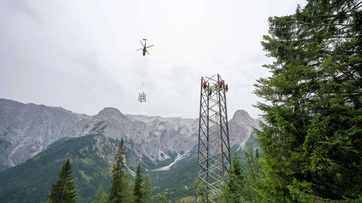 Ein Hubschrauber hebt ein Mastteil ab, während Arbeiter an einem Strommast im Gebirge tätig sind.