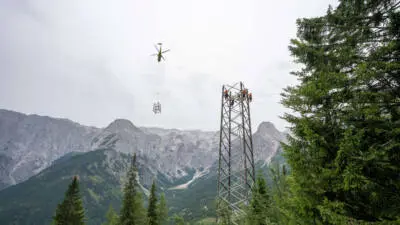 Ein Hubschrauber hebt ein Mastteil ab, während Arbeiter an einem Strommast im Gebirge tätig sind.