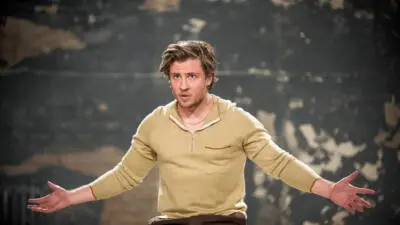 Ein Mann in beigem Pullover sitzt mit ausgestreckten Armen vor einer dunklen Kulisse.